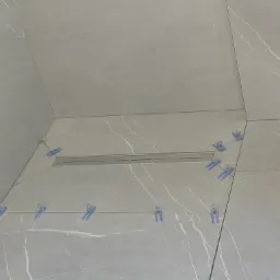 Ułożone szare płytki imitujące marmur, z niebieskimi klipsami poziomującymi i odpływem liniowym. Widok z góry na posadzkę przy ścianie.