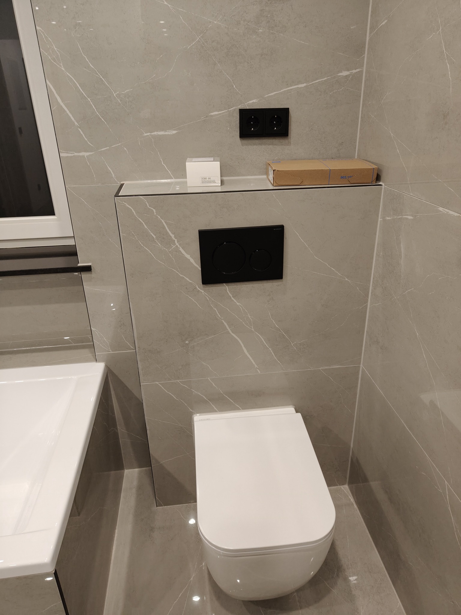 Wykończona łazienka z szarymi płytkami imitującymi marmur, podwieszaną toaletą i umywalką. Widoczny fragment okna i czarne akcenty.