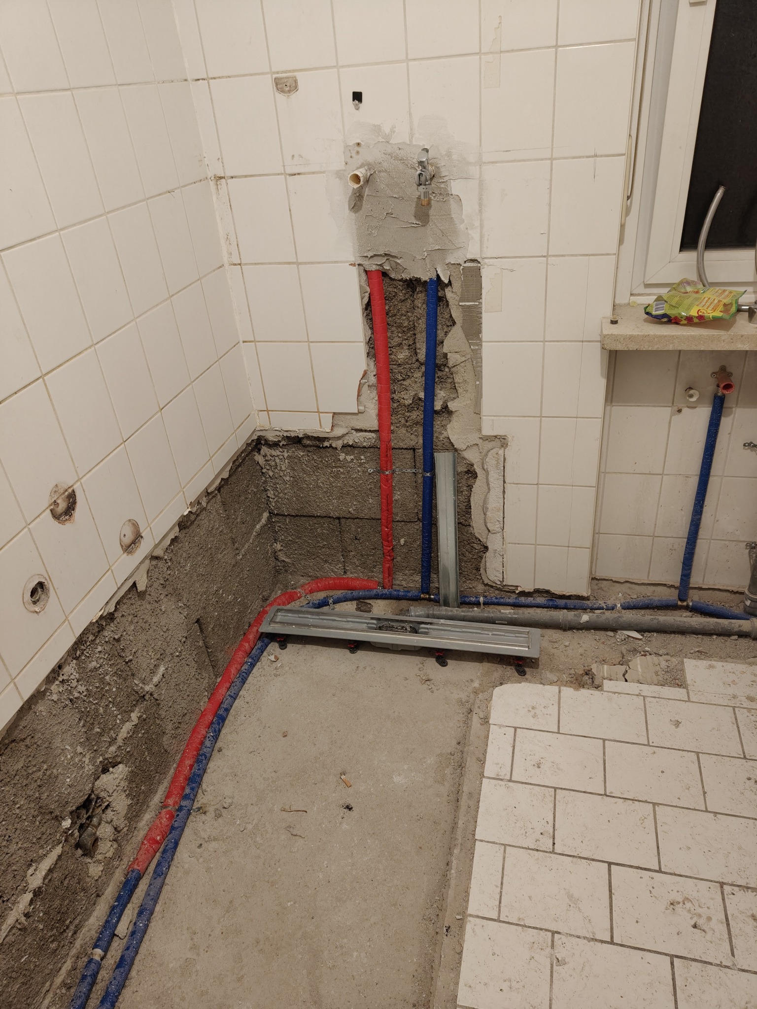 Fragment łazienki w trakcie prac: odsłonięte rury (czerwona i niebieska) na tle białych płytek, częściowo usuniętych, z widocznym gruzem i narzędziami.