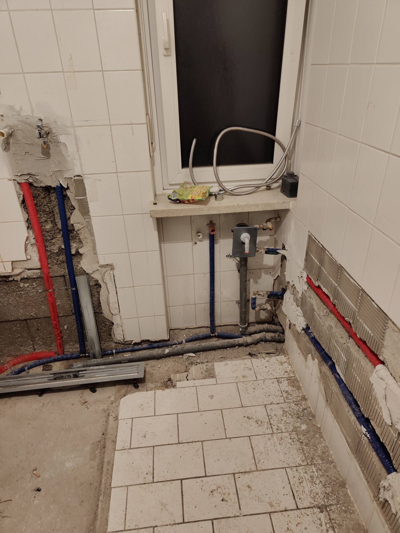 Pionowy kadr łazienki w trakcie prac instalacyjnych: widoczne rury w ścianach, częściowo usunięte płytki i armatura przy oknie. Fragmenty płytek na podłodze.