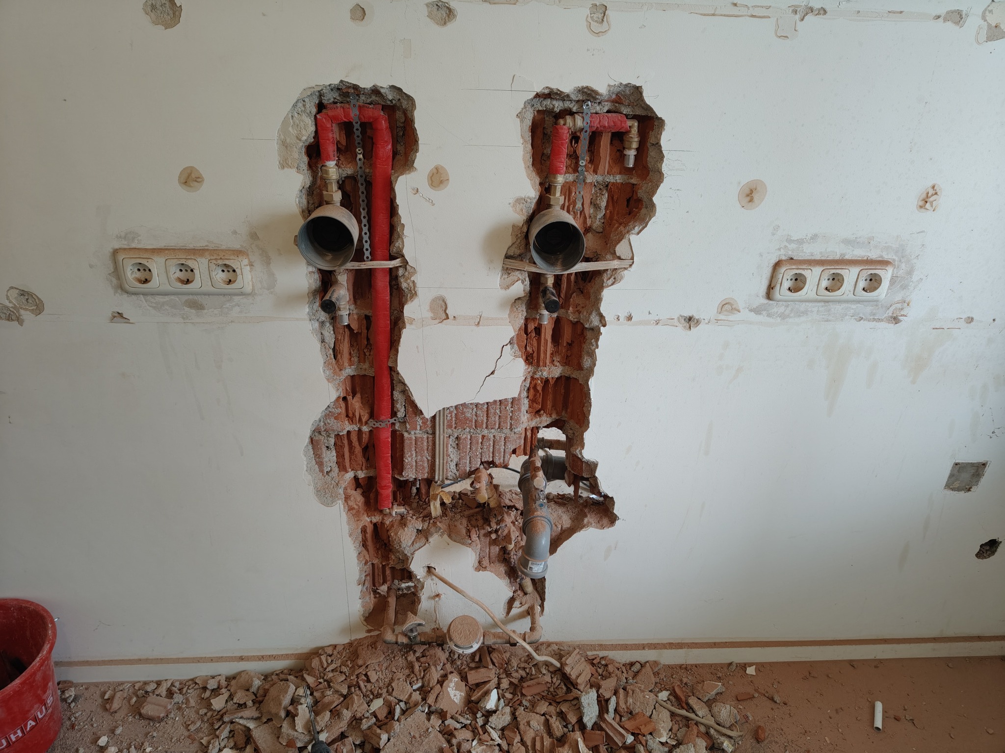 Fragment ściany z odsłoniętą instalacją hydrauliczną i elektryczną, widoczne ślady po kuciu, gruz na podłodze, gniazdka elektryczne. Prace przygotowawcze do remontu.