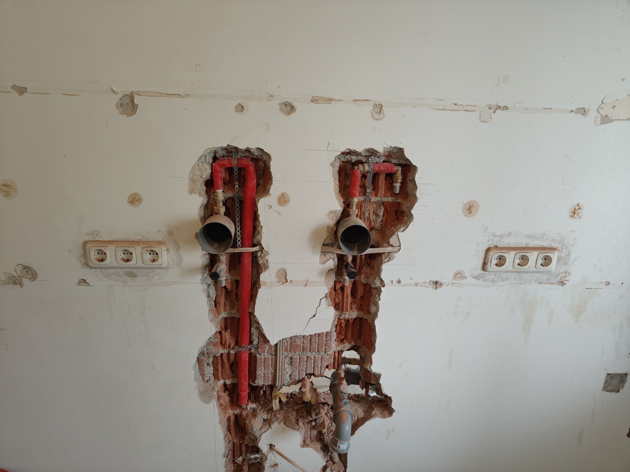 Ściana z widocznymi instalacjami hydraulicznymi w trakcie prac remontowych, z odsłoniętą cegłą i gniazdkami elektrycznymi.