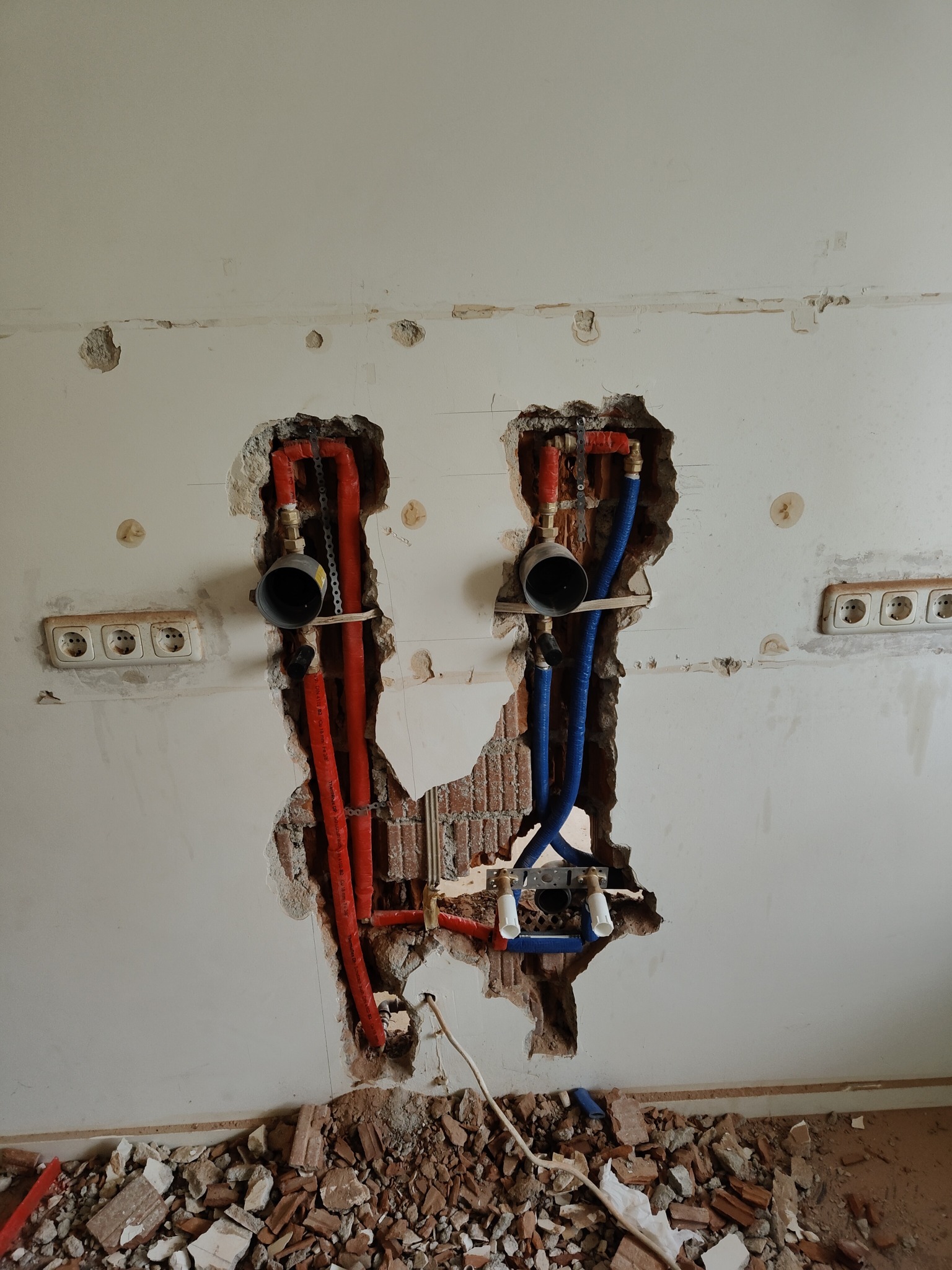 Ściana z odsłoniętymi rurami (czerwone i niebieskie) w dwóch miejscach, widoczne gniazdka elektryczne i gruz na podłodze. Etap prac instalacyjnych wewnątrz budynku.