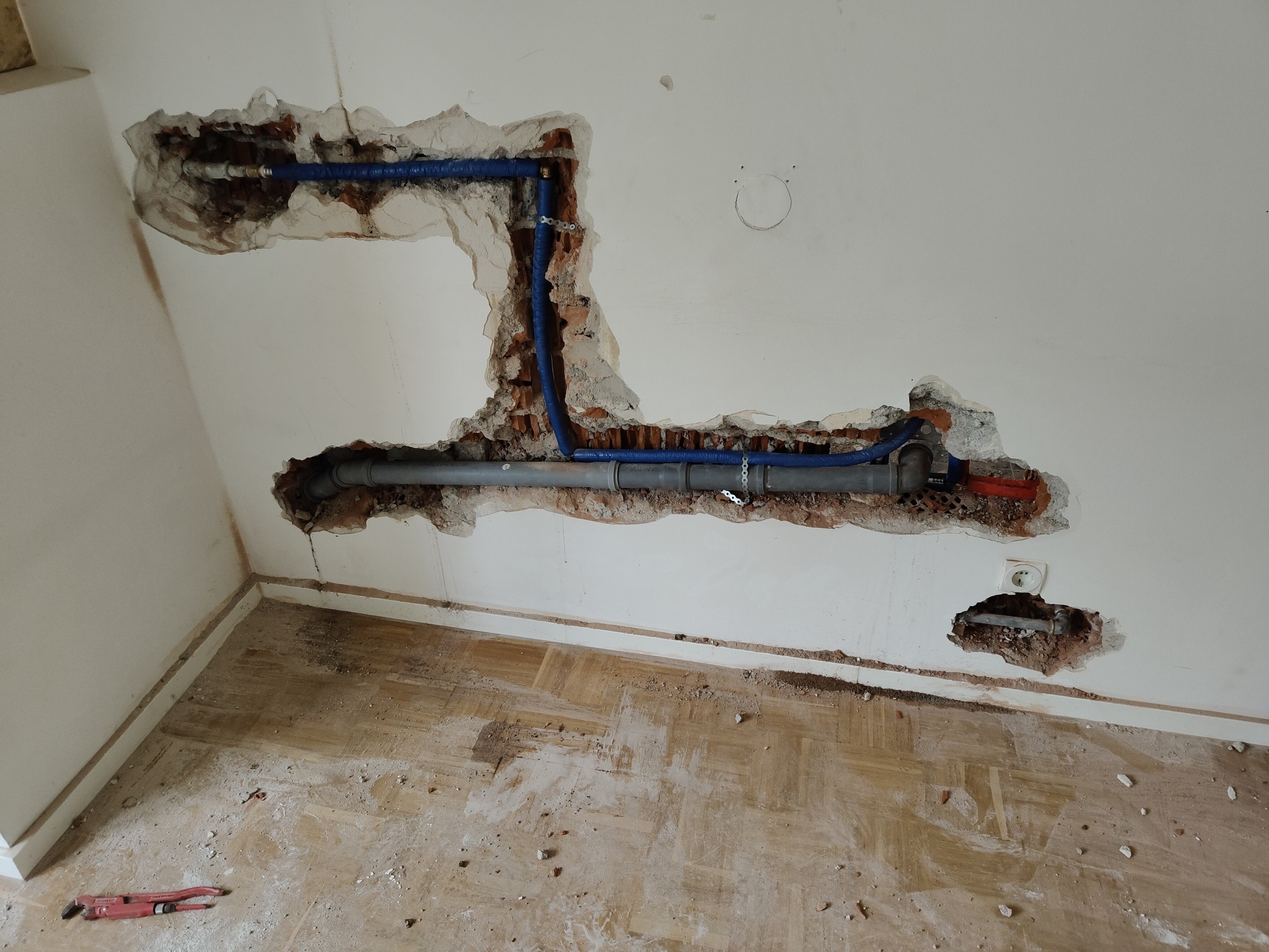 Ściana w trakcie prac instalacyjnych: widoczne niebieskie rury prowadzone w wykuciach, surowy stan, narzędzie na podłodze. Układanie instalacji wewnątrz budynku.