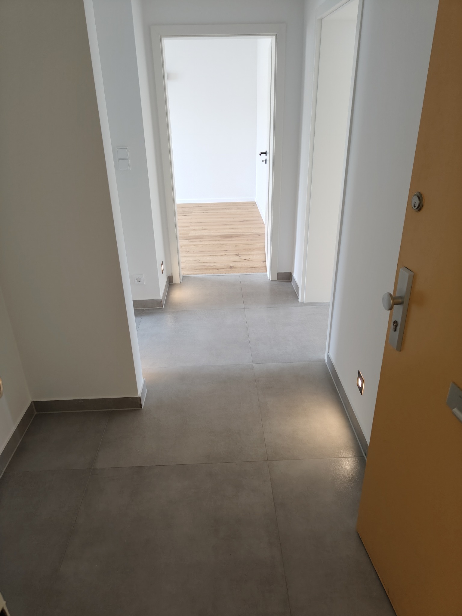 Nowoczesny korytarz z szarymi płytkami podłogowymi i minimalistycznym oświetleniem. W tle widoczne otwarte drzwi do pokoju z drewnianą podłogą.