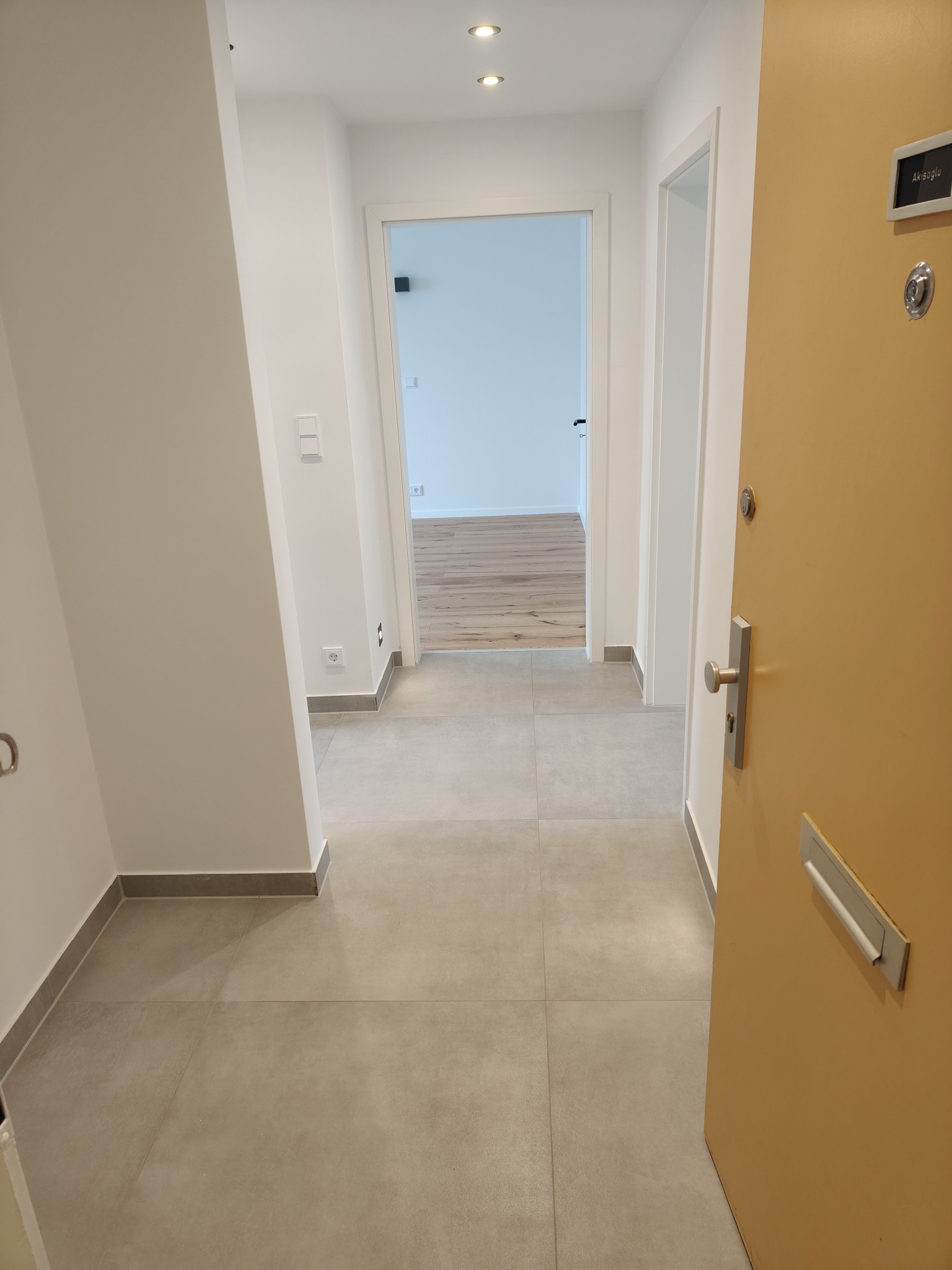 Jasne wnętrze mieszkania po wykończeniu: szare płytki podłogowe, białe ściany, widoczne drzwi wejściowe i otwarte drzwi do kolejnych pomieszczeń z drewnianą podłogą.