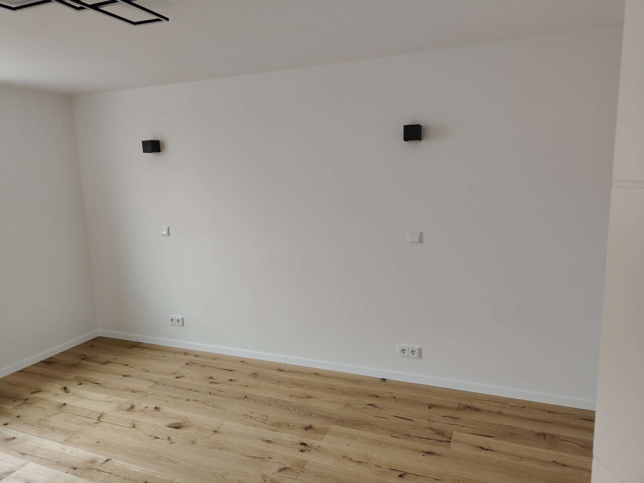 Jasne panele podłogowe w pokoju z białymi ścianami i minimalistycznymi, czarnymi lampami ściennymi. Widoczne gniazdka i listwy przypodłogowe.