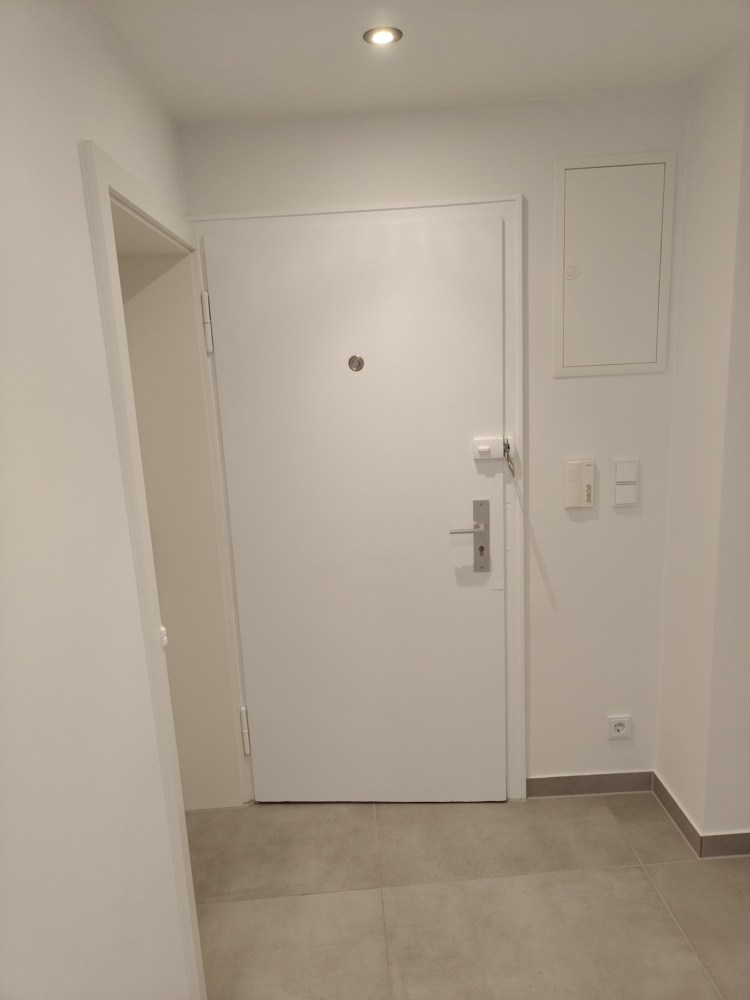 Białe drzwi wejściowe z wizjerem i zamkiem, widok na korytarz z płytkami podłogowymi. Świeże, minimalistyczne wykończenie wnętrza.