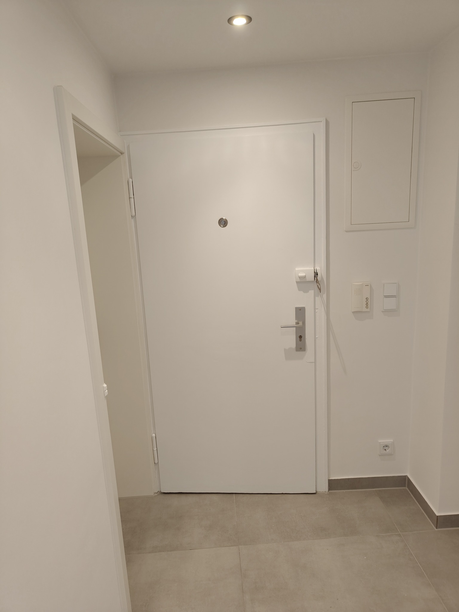 Białe drzwi wejściowe z wizjerem i srebrną klamką w jasnym korytarzu z szarą podłogą. Widoczny fragment otwartych drzwi po lewej i skrzynka elektryczna po prawej.