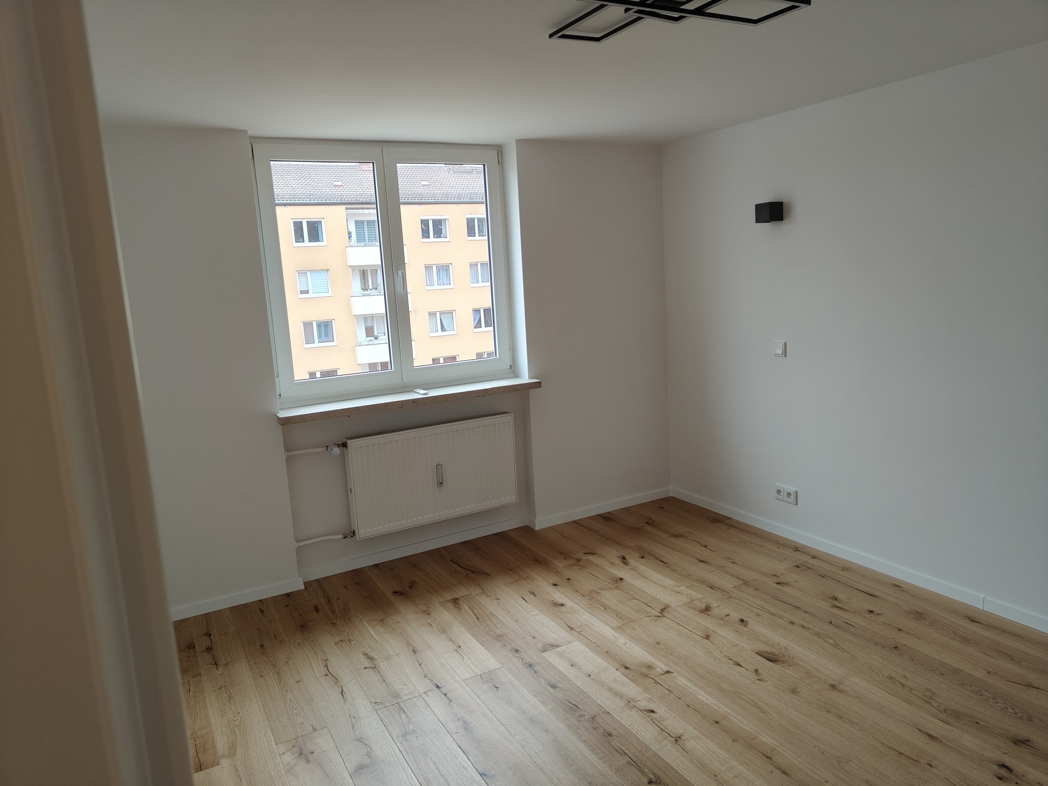 Jasne panele podłogowe w pustym pokoju z białymi ścianami, okno z widokiem na budynek mieszkalny, nowoczesna lampa sufitowa i grzejnik pod oknem.