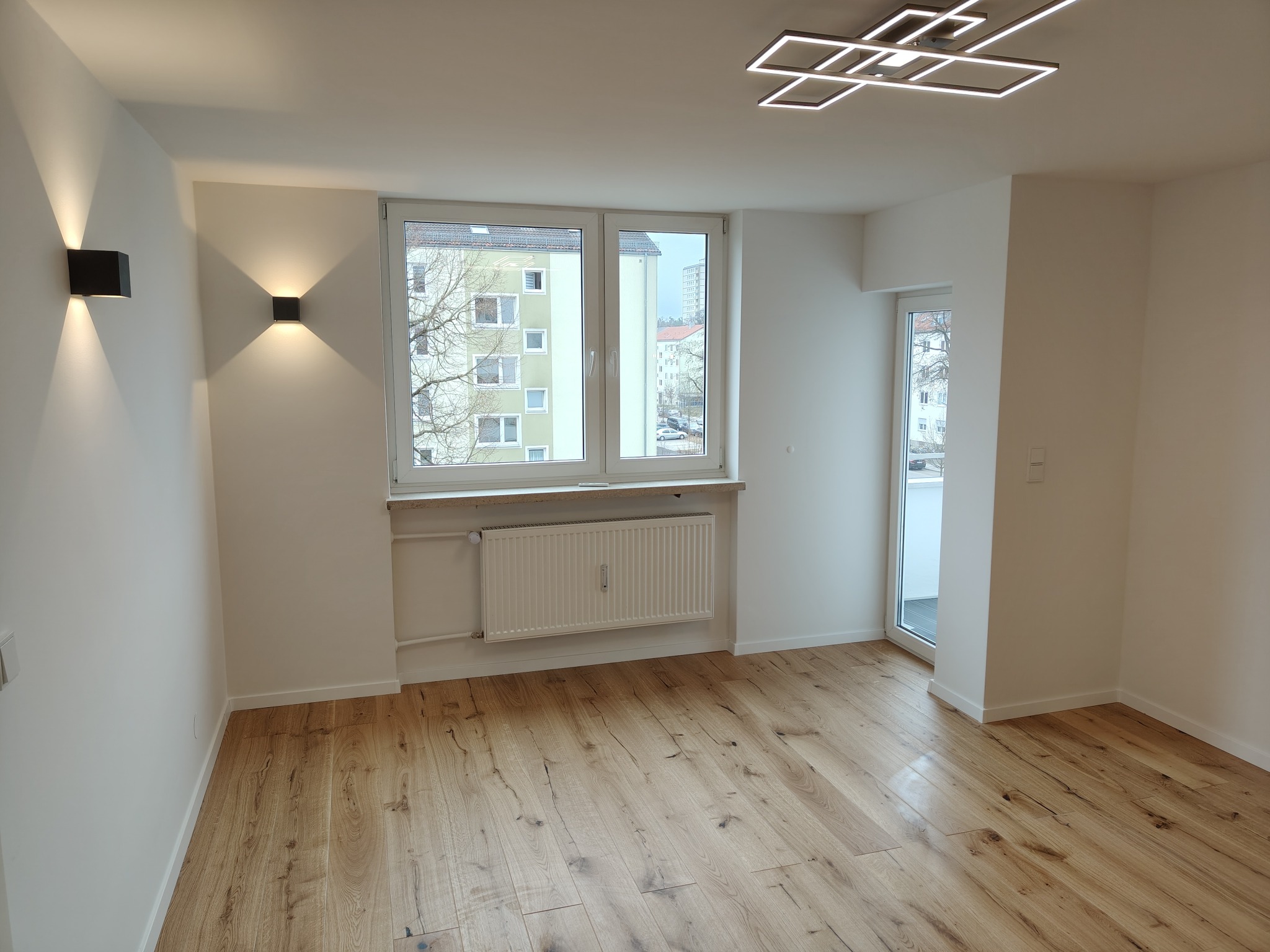 Jasne, puste wnętrze z drewnianą podłogą, białymi ścianami, oknem z widokiem na budynek i nowoczesnym oświetleniem sufitowym. Minimalistyczny design.