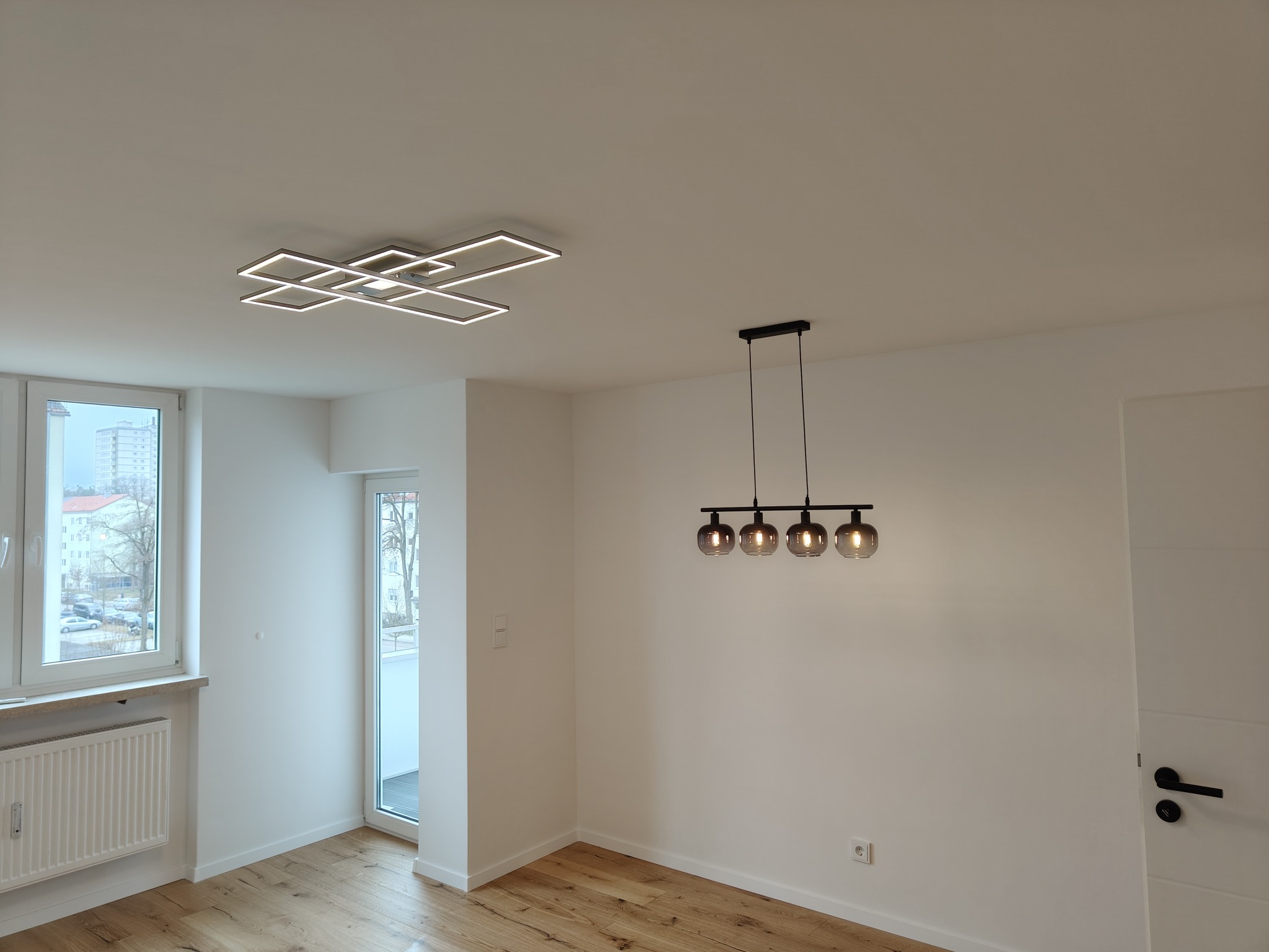 Jasne, przestronne wnętrze po odnowieniu w Wolnzach: parkiet, białe ściany, nowoczesne lampy sufitowe i okno z widokiem na miasto. Minimalistyczny design.