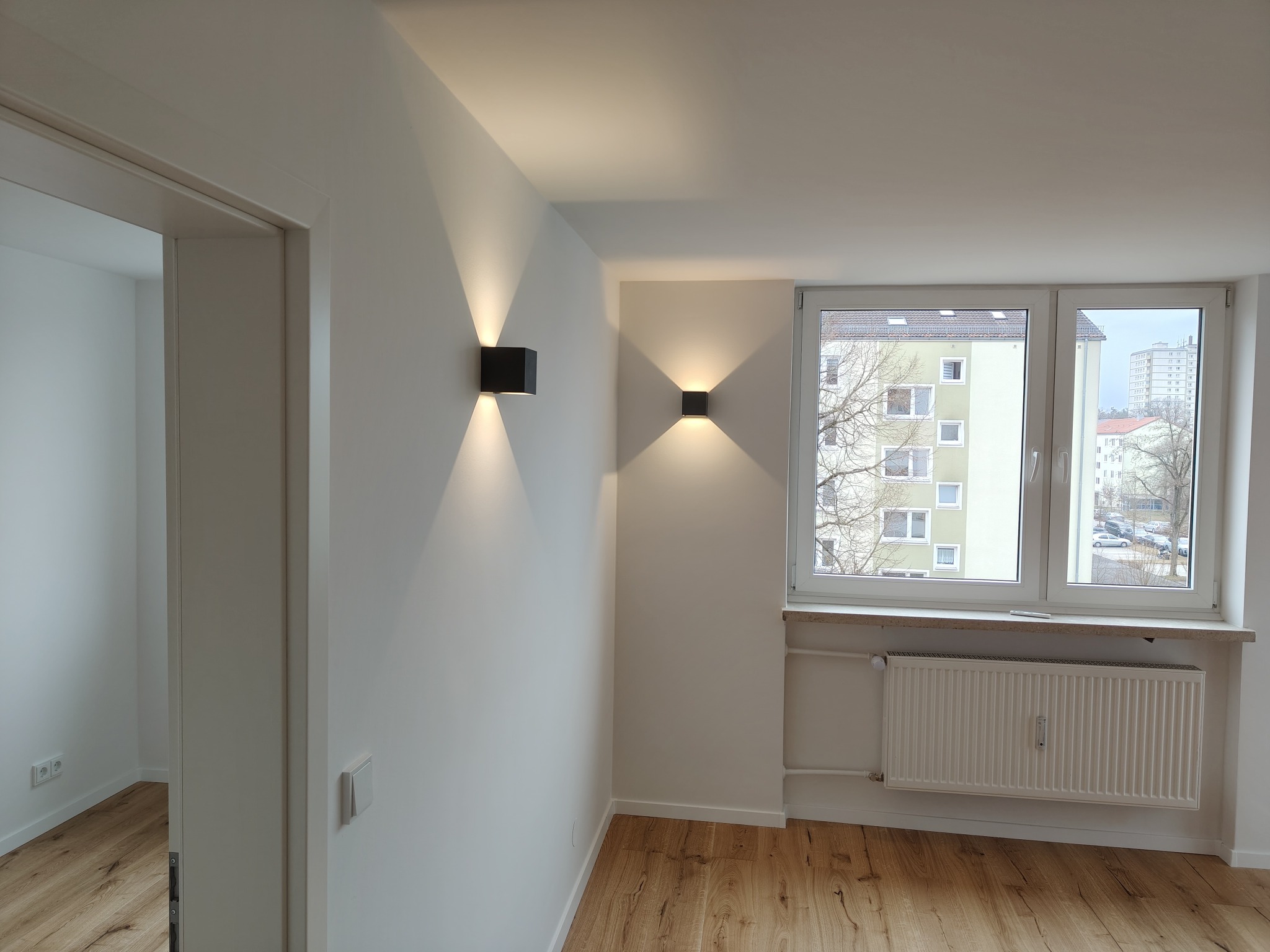 Wnętrze po remoncie: parkiet, białe ściany z nowoczesnymi lampami, okno z widokiem na miasto. Minimalistyczny design, jasne kolory, czyste linie.