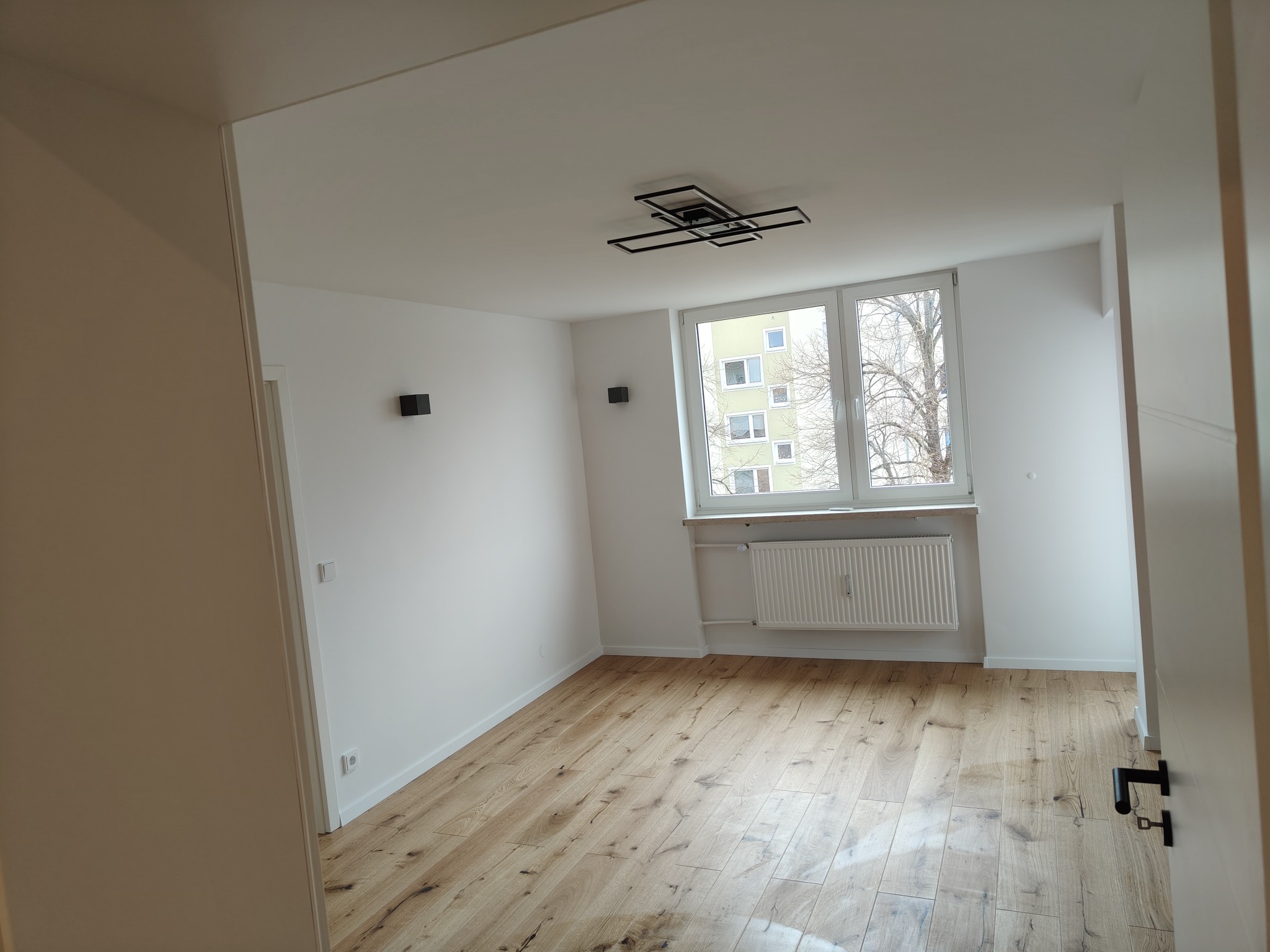 Jasne wnętrze z drewnianą podłogą, białymi ścianami i nowoczesną lampą sufitową. Widok przez otwarte drzwi na pokój z oknem i grzejnikiem.