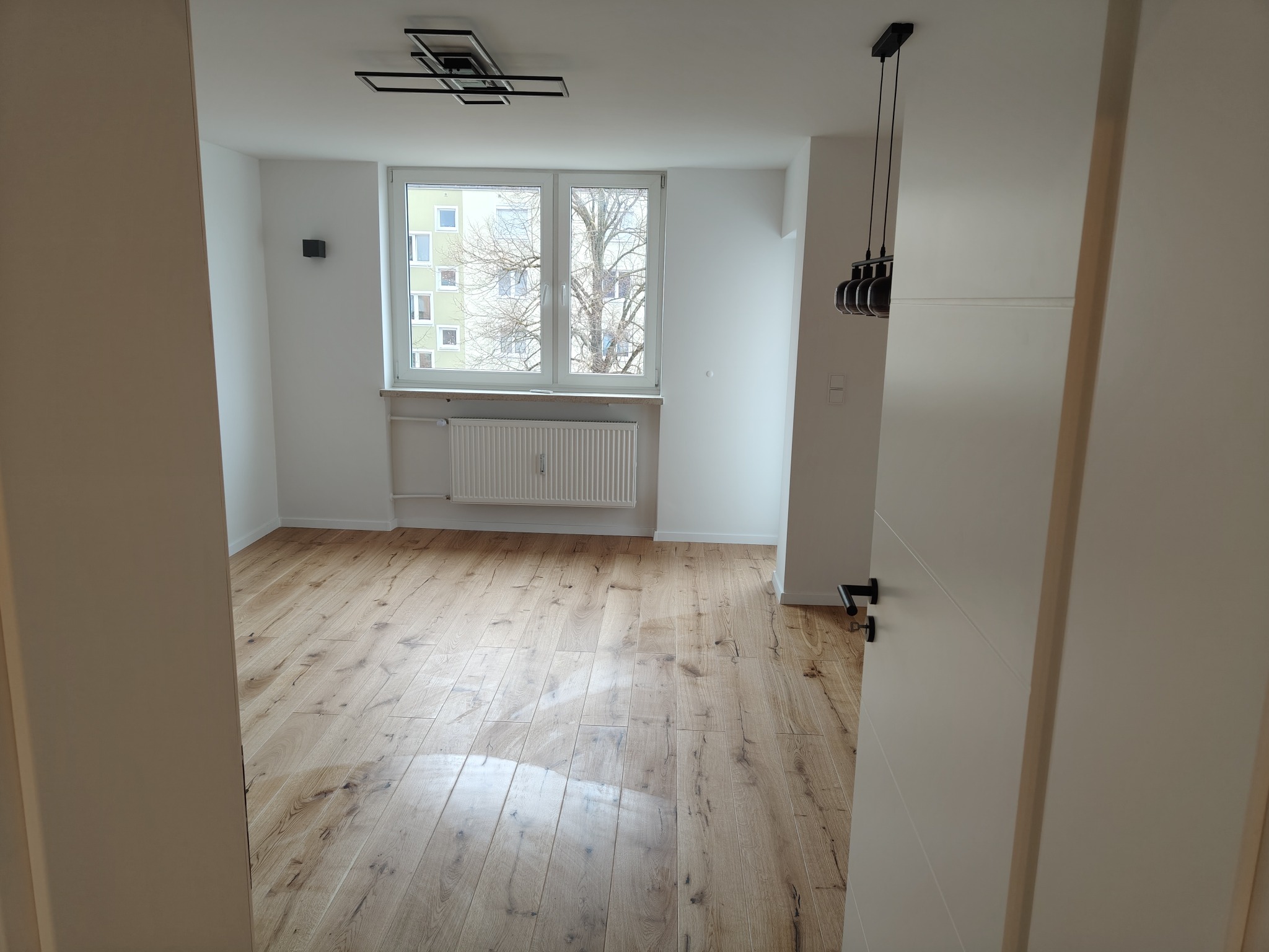 Jasne, drewniane panele podłogowe w wyremontowanym pokoju z białymi ścianami, widokiem z okna na budynek i drzewa oraz nowoczesnym oświetleniem.