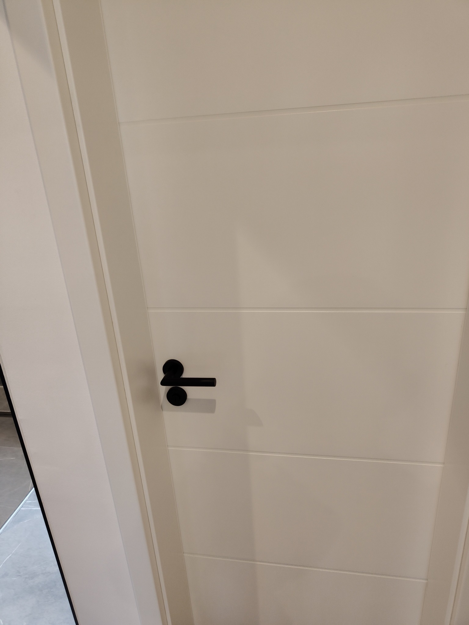 Białe drzwi z czarną klamką i subtelnymi poziomymi frezami, widoczne w kadrze od dołu, z fragmentem podłogi w tle.