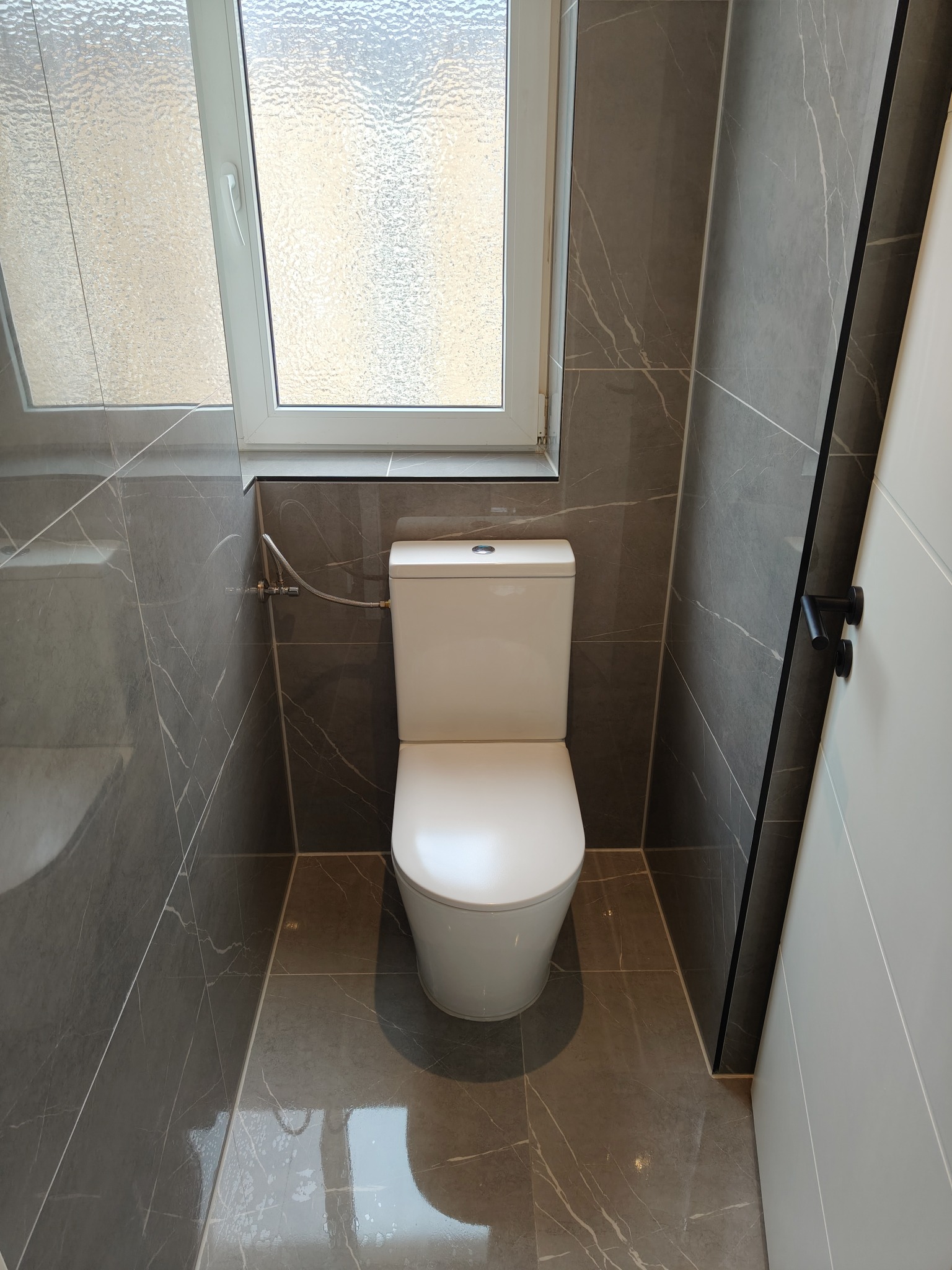 Nowoczesna toaleta z szarymi płytkami imitującymi marmur, białą armaturą i oknem z matową szybą. Widok z góry na posadzkę z połyskiem.