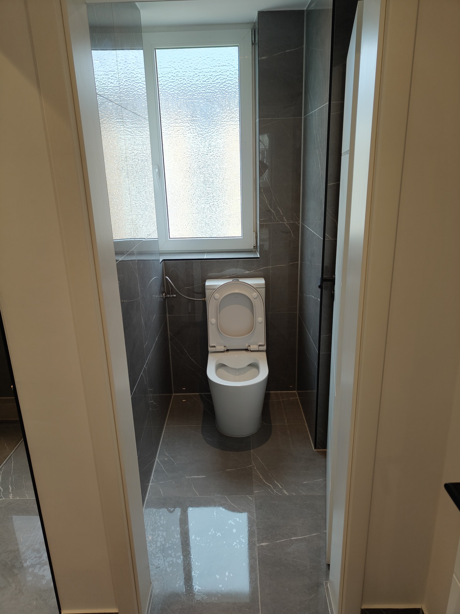 Nowoczesna toaleta z szarymi płytkami imitującymi marmur, widoczna przez białe futryny drzwi. Okno z matową szybą w tle. Czyste i jasne wnętrze.