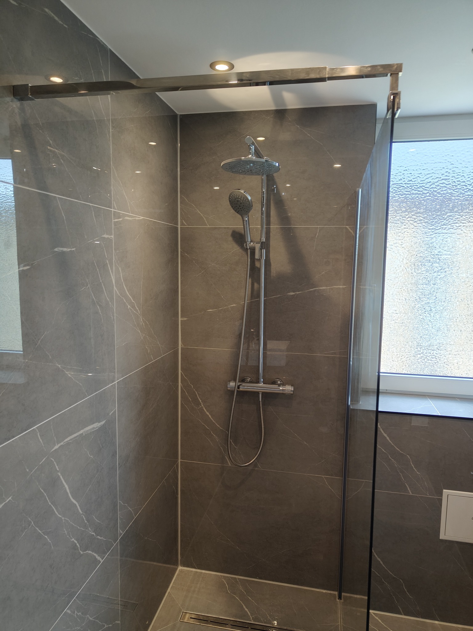 Nowoczesna kabina prysznicowa z deszczownicą i szklaną ścianką, wykończona szarymi płytkami imitującymi marmur, z odpływem liniowym w podłodze.