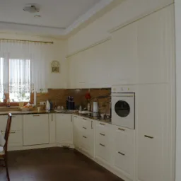 Jasna kuchnia w klasycznym stylu z kremowymi szafkami, marmurowym blatem i brązową podłogą; okno z koronkowymi firankami i stół z krzesłami w tle.