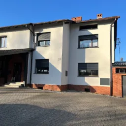 Dwukondygnacyjny dom z ocieploną elewacją w odcieniach bieli, z ciemnymi roletami zewnętrznymi i ceglaną podmurówką, numer domu 18, widoczny fragment podjazdu z kostki brukowej i boczna dobudówka...