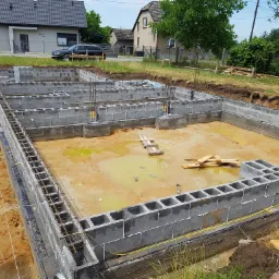 Wykopane fundamenty pod budynek mieszkalny, widoczne bloczki betonowe tworzące zarys ścian, zbrojenie pionowe i poziome, żółta woda gruntowa w wykopie, w tle dom i samochód.