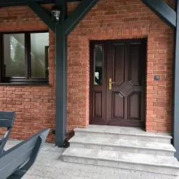 Wejście do domu z ceglaną elewacją, brązowymi drzwiami z subtelnymi zdobieniami, szarymi schodami z płytek i drewnianą konstrukcją wspornikową pomalowaną na szaro.