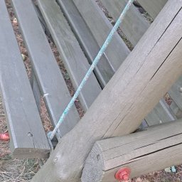 Usługi ogrodowe Zielony Majster Paweł Romaniszyn - Drewniana huśtawka ogrodowa w odcieniach zieleni, zawieszona na linach, z widocznymi czerwonymi śrubami montażowymi i opadłymi owocami na trawie.