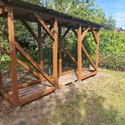 Drewniana konstrukcja z zadaszeniem w ogrodzie, prawdopodobnie przeznaczona na składowanie drewna, z przygotowanym miejscem na palety, otoczona zielenią i gałęziami.