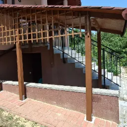 Zadaszenie schodów zewnętrznych z drewnianą kratownicą i brązowymi słupami, widok na kamienną ścieżkę i zieleń ogrodu w tle.