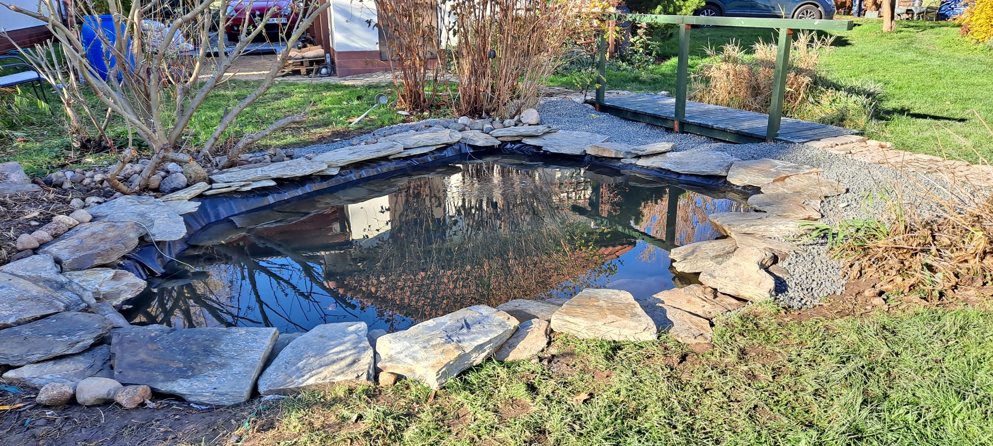 Oczko wodne z kamienną obudową i małym drewnianym mostkiem w ogrodzie, woda odbija niebo, a brzeg wyłożony jest ozdobnym żwirem.