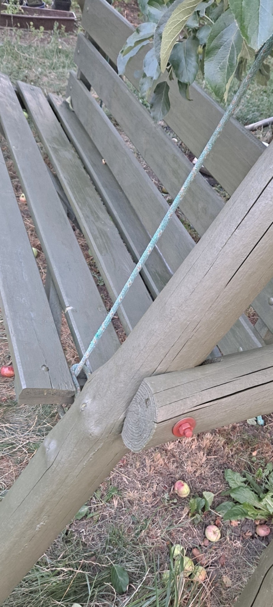 Drewniana huśtawka ogrodowa w odcieniach zieleni, zawieszona na linach, z widocznymi czerwonymi śrubami montażowymi i opadłymi owocami na trawie.