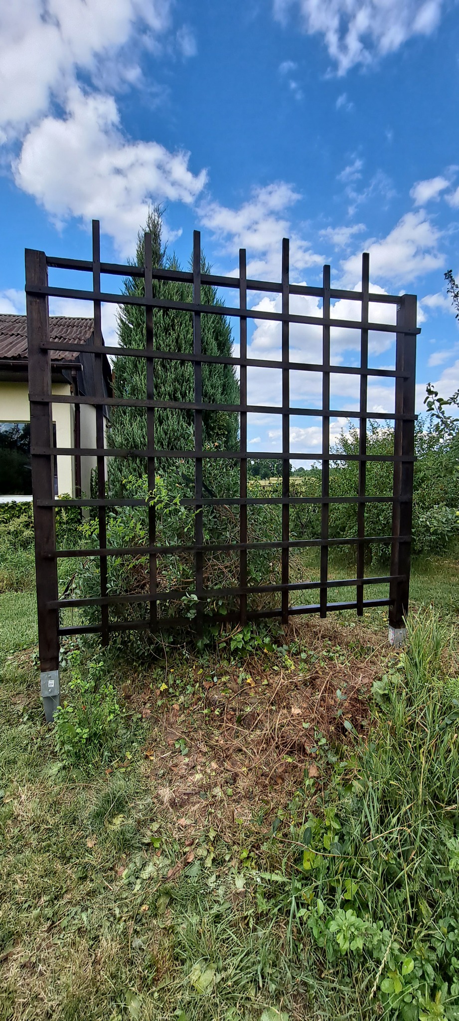 Ciemna, drewniana pergola ogrodowa w kratkę, z widocznym montażem do podłoża, na tle zieleni i błękitnego nieba z chmurami.