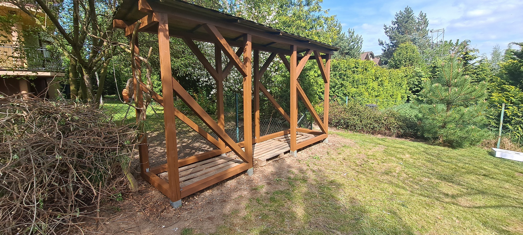 Drewniana konstrukcja z zadaszeniem w ogrodzie, prawdopodobnie przeznaczona na składowanie drewna, z przygotowanym miejscem na palety, otoczona zielenią i gałęziami.