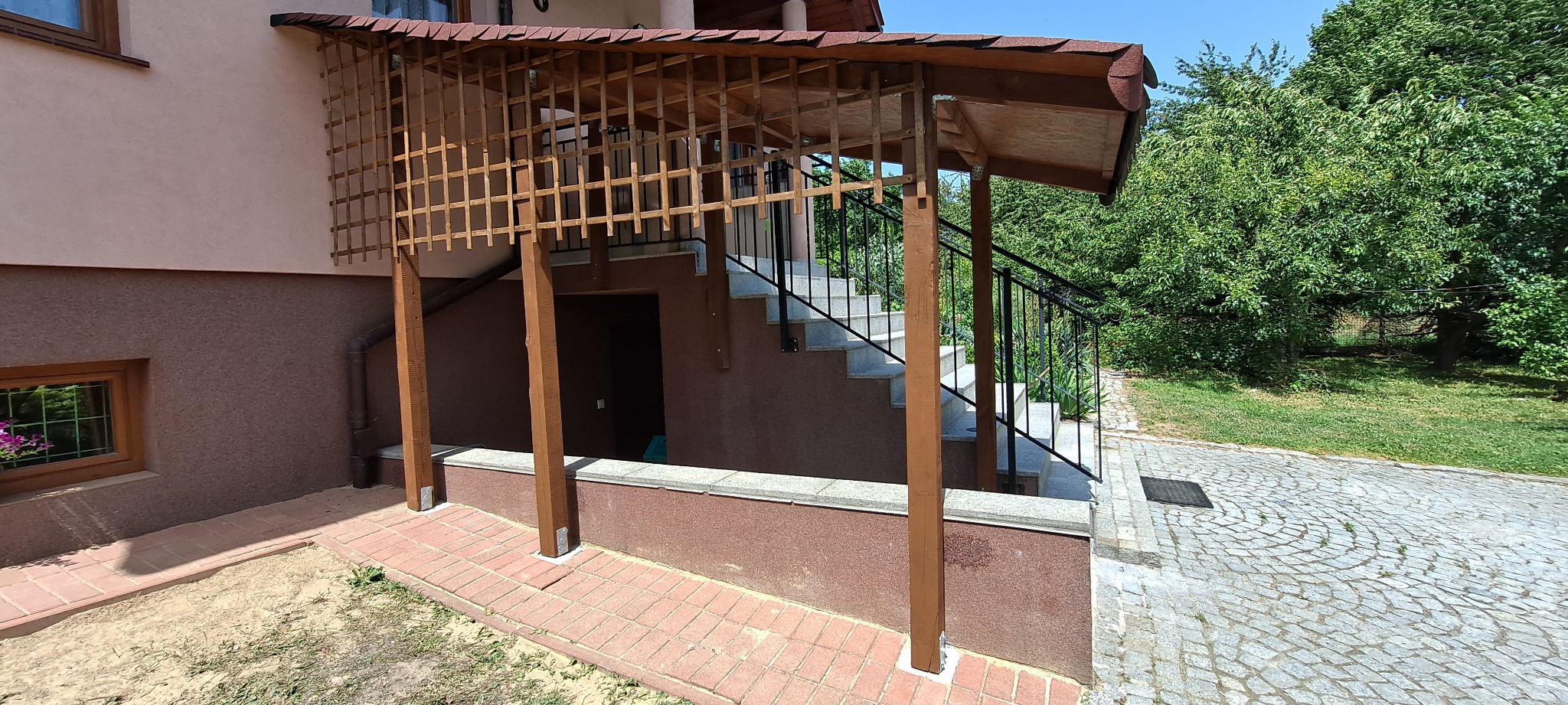 Zadaszenie schodów zewnętrznych z drewnianą kratownicą i brązowymi słupami, widok na kamienną ścieżkę i zieleń ogrodu w tle.
