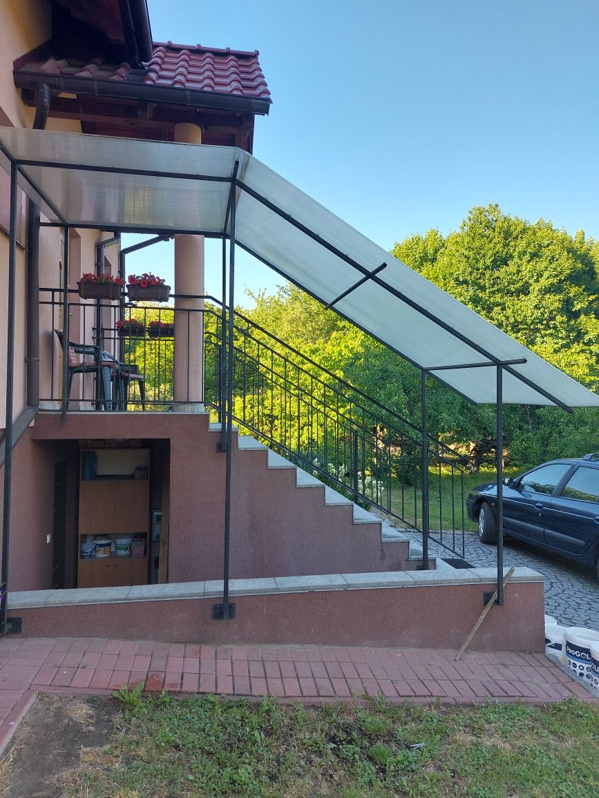 Zadaszone schody wejściowe do domu z czarną metalową balustradą, donicami z czerwonymi kwiatami i widocznym samochodem w tle.