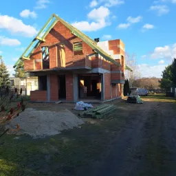 Surowy, dwukondygnacyjny dom murowany w trakcie budowy, z widocznym szkieletem dachu i ścianami z czerwonej cegły, na tle błękitnego nieba z chmurami.