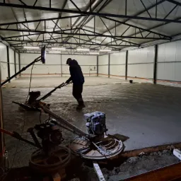 Wyrównywanie wylewki betonowej w hali przemysłowej za pomocą zacieraczek mechanicznych. Widoczne ślady pracy maszyn na powierzchni betonu i konstrukcja stalowa dachu.