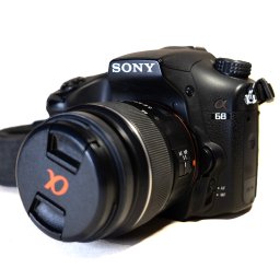 Art Foto Klaudia Ziomko - Czarny aparat Sony Alpha 68 z założoną osłoną obiektywu z pomarańczowym logo Alpha na białym tle, ustawiony pod kątem, widoczny pasek.