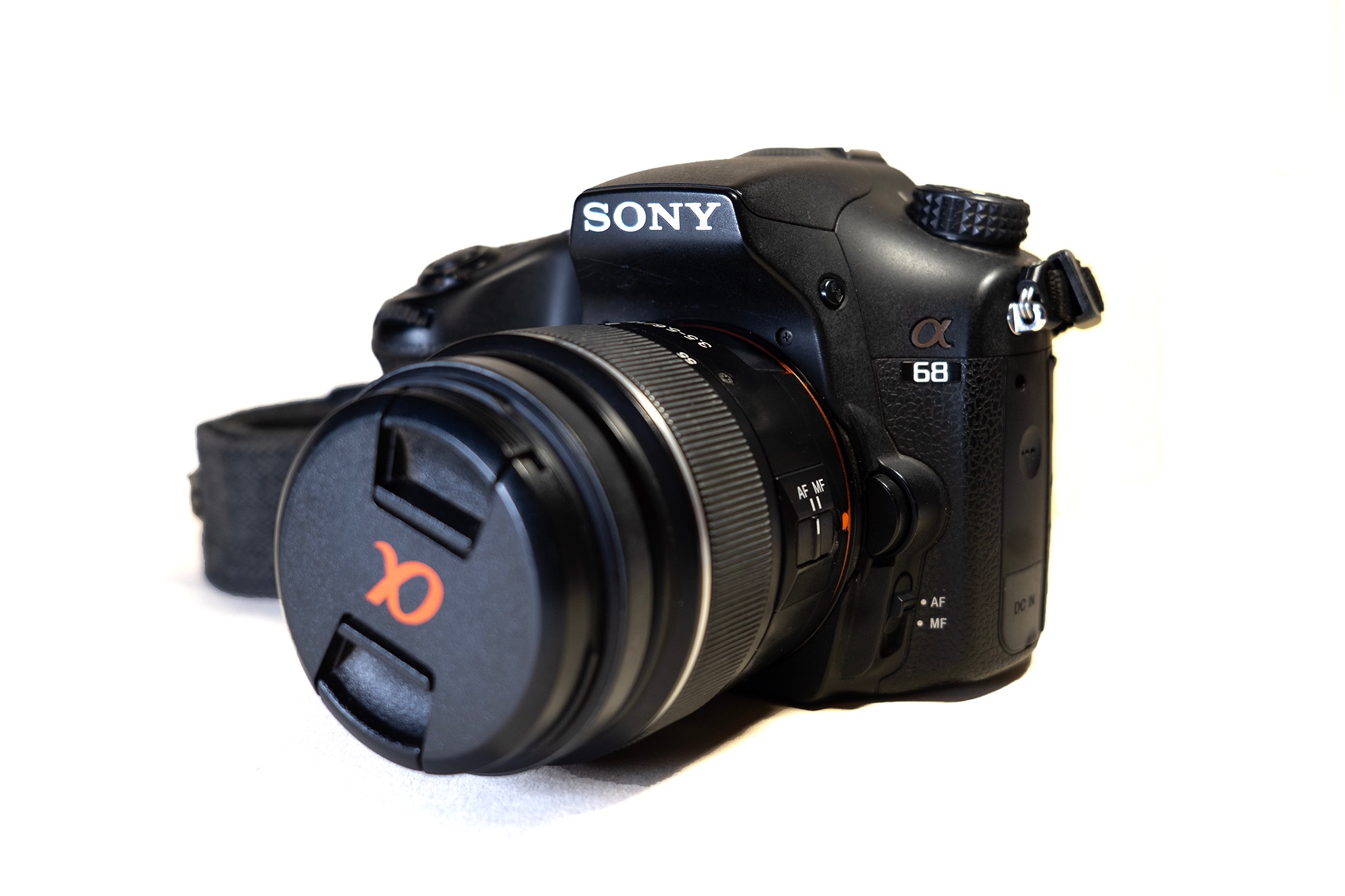 Czarny aparat Sony Alpha 68 z założoną osłoną obiektywu z pomarańczowym logo Alpha na białym tle, ustawiony pod kątem, widoczny pasek.