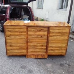 Zakład Stolarski - Odrestaurowana komoda z forniru w stylu art deco, umieszczona na tle miejskiej ulicy z otwartym bagażnikiem samochodu.