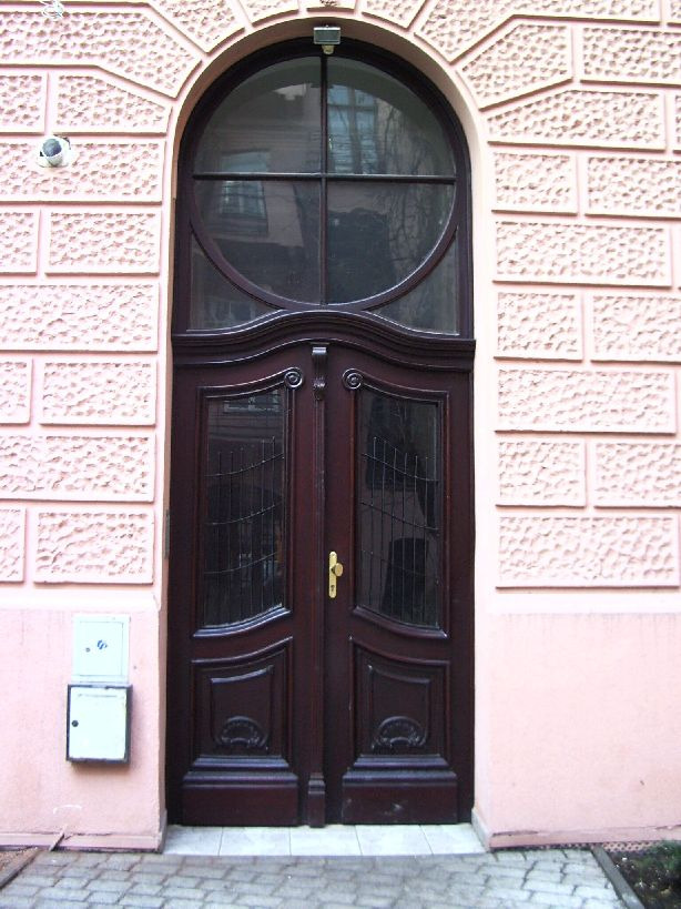 Odnowione, dwuskrzydłowe, ciemnobrązowe drzwi wejściowe z okrągłym oknem nadświetlnym w kamienicy z różową elewacją o fakturze bloków.