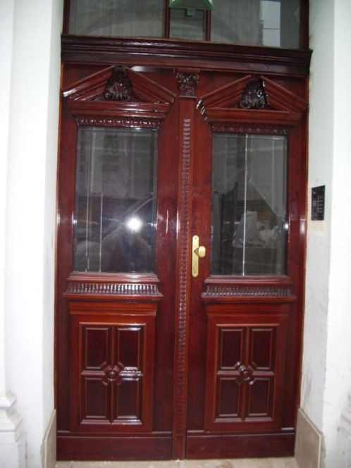 Odrestaurowane dwuskrzydłowe drzwi wejściowe z ciemnego drewna, z dekoracyjnymi rzeźbieniami i przeszkleniami, widziane z bliska, z widoczną złotą klamką i domofonem po prawej stronie.