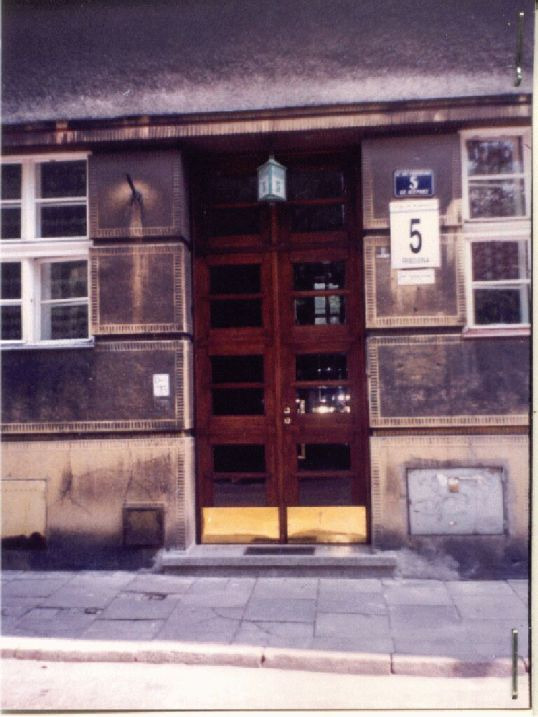 Frontowe wejście do budynku z drewnianymi drzwiami z szybami, numerem 5 na ścianie i ozdobną latarnią wiszącą nad wejściem.