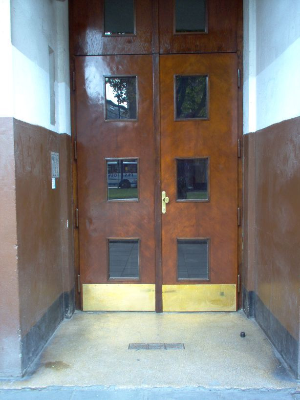 Odrestaurowane, dwuskrzydłowe, drewniane drzwi wejściowe z małymi, kwadratowymi okienkami, zabezpieczone u dołu złotą, metalową płytą, osadzone w brązowo-białym portalu.