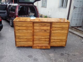 Odrestaurowana komoda z forniru w stylu art deco, umieszczona na tle miejskiej ulicy z otwartym bagażnikiem samochodu.