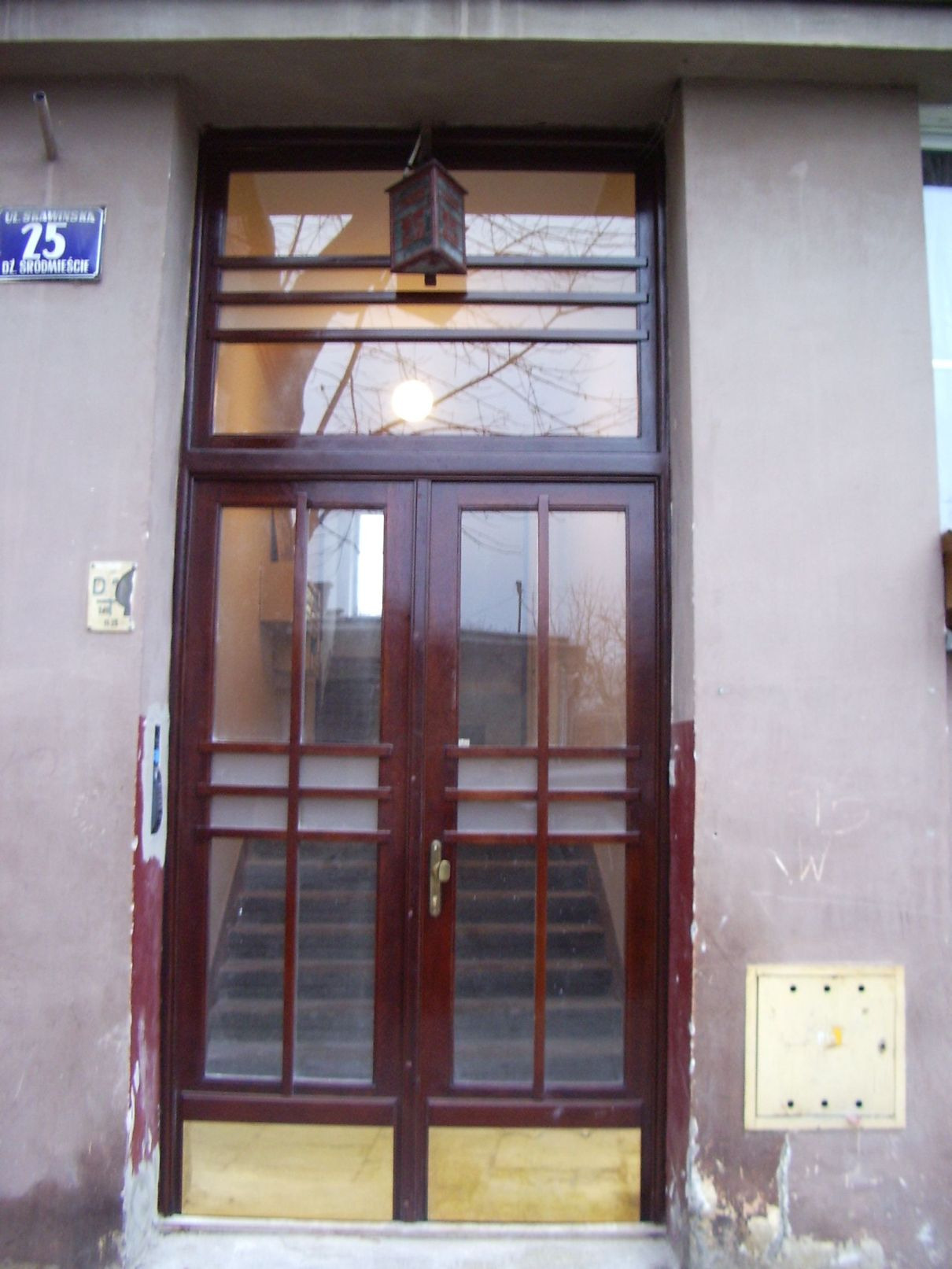 Brązowe, przeszklone drzwi wejściowe do budynku z widoczną klatką schodową wewnątrz, oświetlone wiszącą lampą i odblaskiem żarówki, z tabliczką adresową 'Ul. Bramińska 25' po lewej stronie.