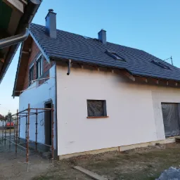 Dom jednorodzinny w trakcie ocieplania elewacji, widoczne rusztowania, częściowo otynkowana ściana, okna zabezpieczone folią, fragment ściany z widocznym ociepleniem styropianem.