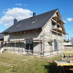Dom jednorodzinny w trakcie ocieplania, widoczne rusztowania, nieotynkowana elewacja w kolorze szarym, dach pokryty ciemną dachówką, błękitne niebo z chmurami w tle.