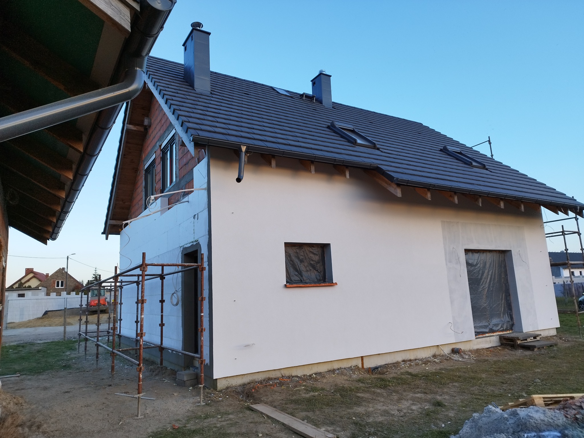 Dom jednorodzinny w trakcie ocieplania elewacji, widoczne rusztowania, częściowo otynkowana ściana, okna zabezpieczone folią, fragment ściany z widocznym ociepleniem styropianem.